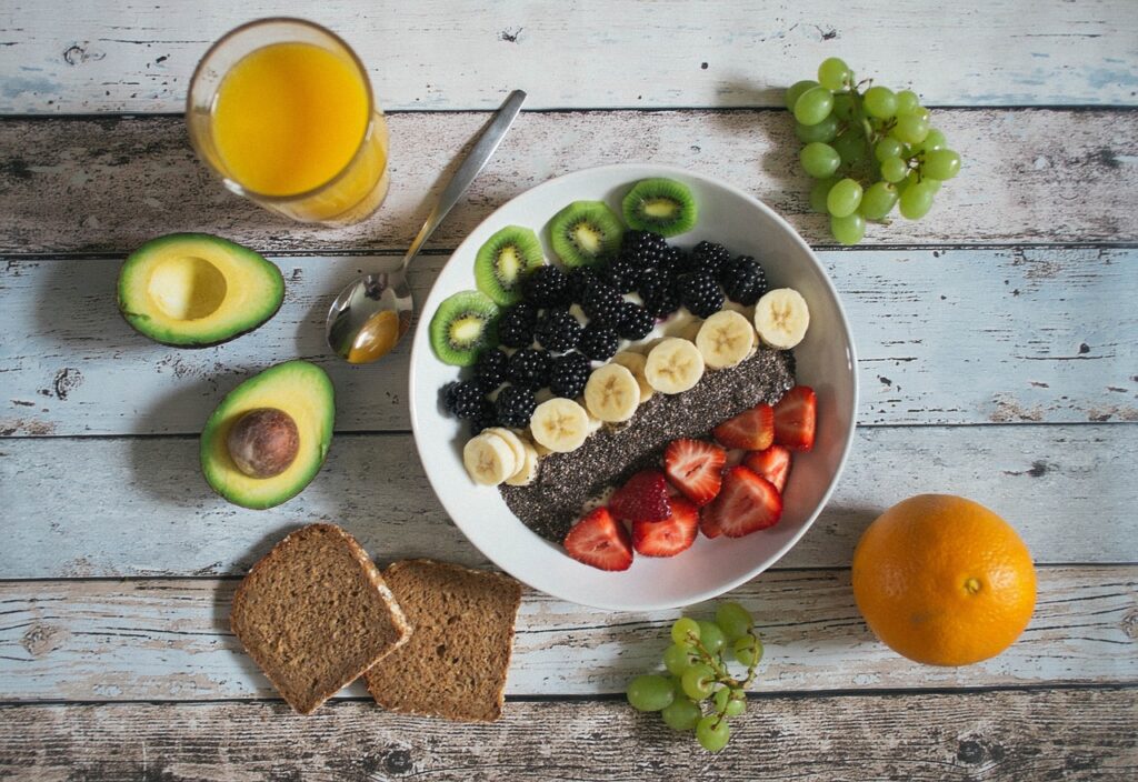 A nutritious breakfast bowl containing banana slices, blueberries, pecans, and cereal, placed on a wooden table with a spoon, a notebook, white flowers, and additional bowls of fruits and nuts.