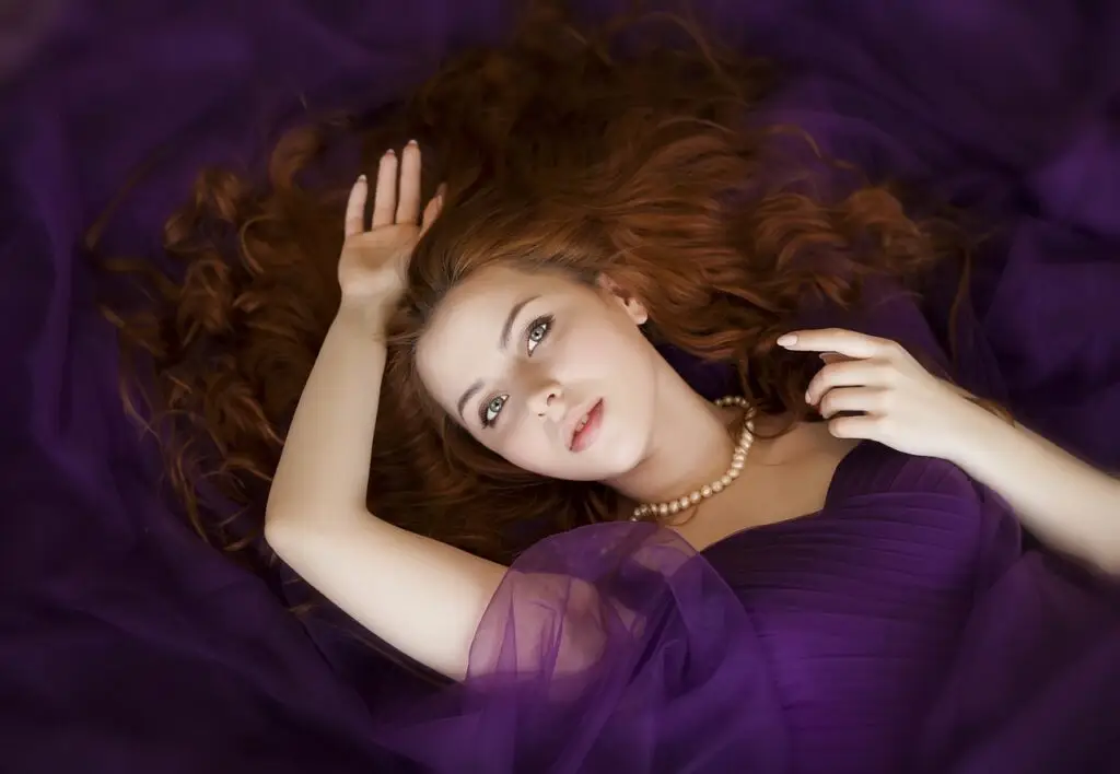 Beautiful woman with radiant skin lying on purple fabric, wearing a pearl necklace.